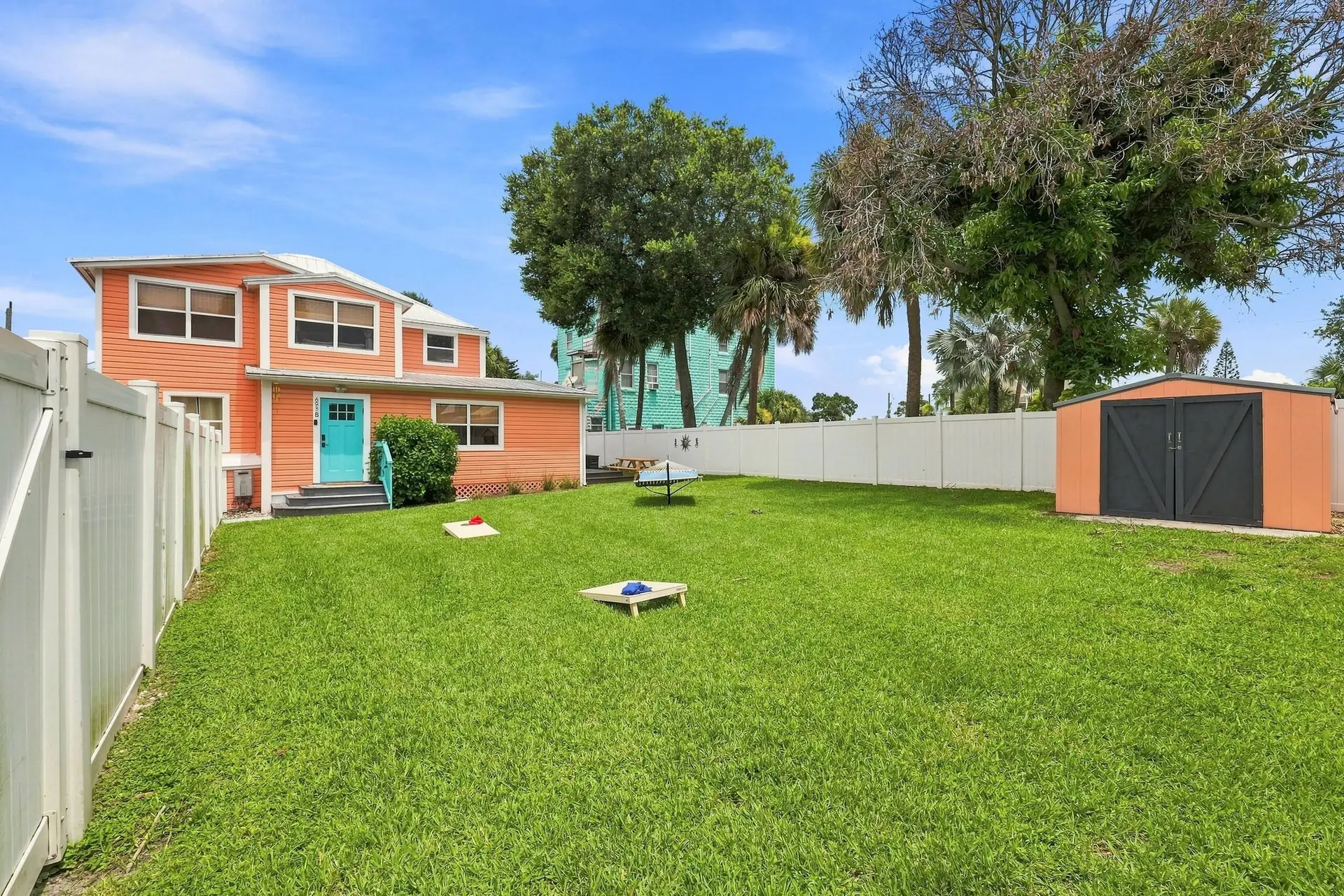 Large fenced backyard with corn-hole boards set up on the grass, a hammock, a storage shed and palm trees