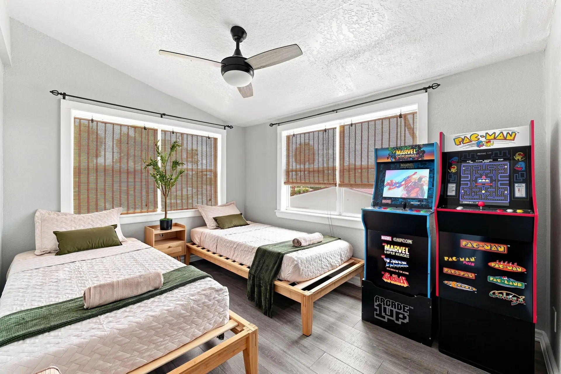 Bedroom with two XL twin beds on light wood platform frames, olive green throws, and two stand-up arcade cabinets in the corner
