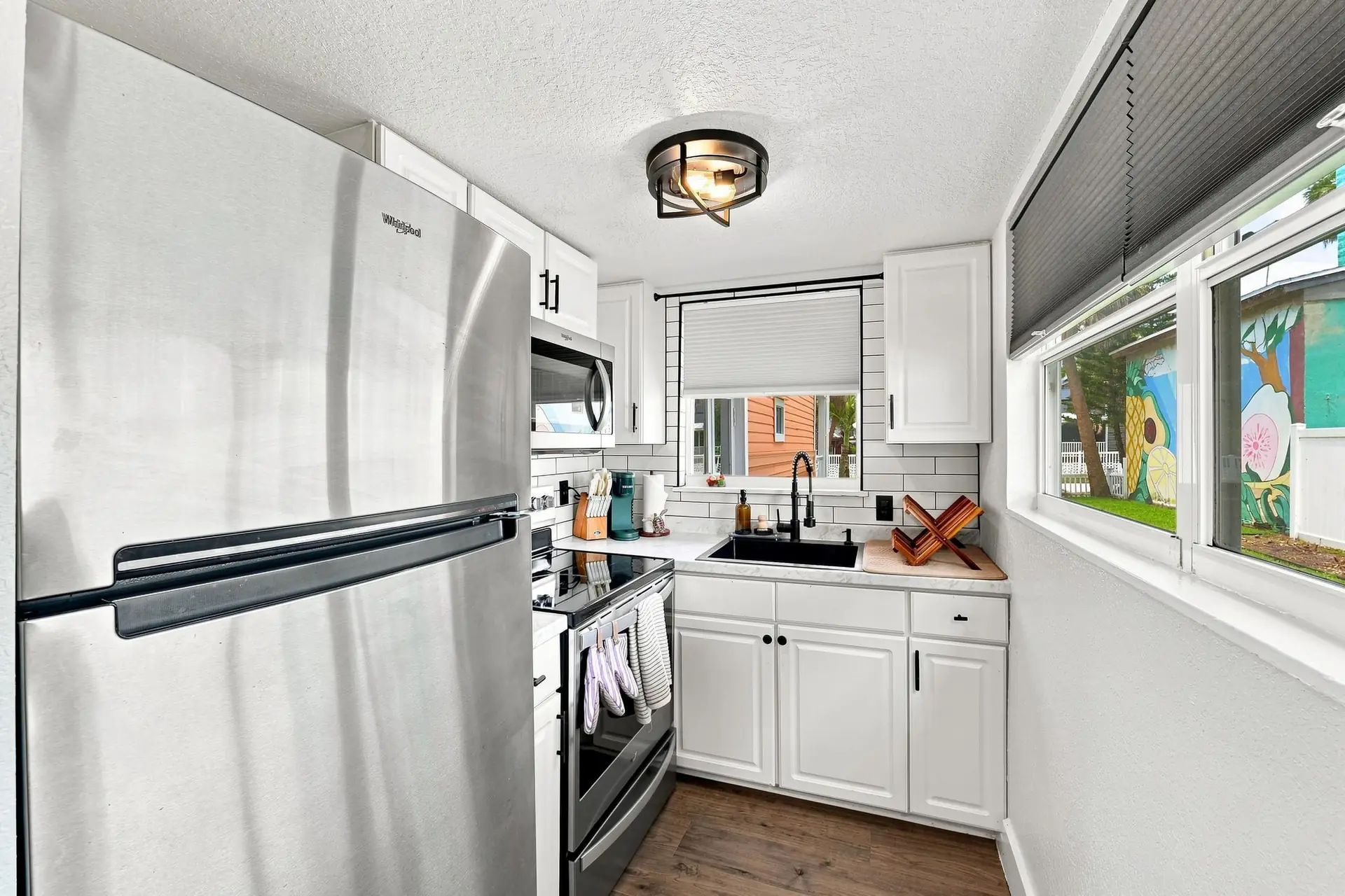 Galley kitchen with a full-size stainless refrigerator, range and coffee maker, with windows looking out onto a painted neighborhood mural